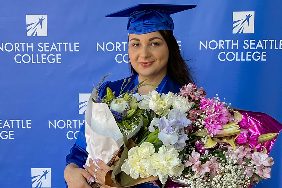 female in blue cap and gown holding flowers in front of a blue backdrop with North Seattle College logo