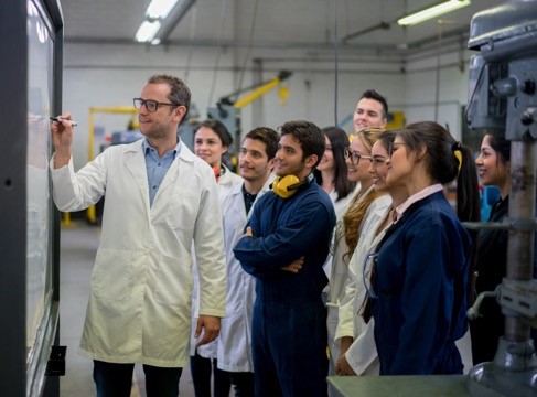 male in white lab coat drawing on white board with students standing next to him