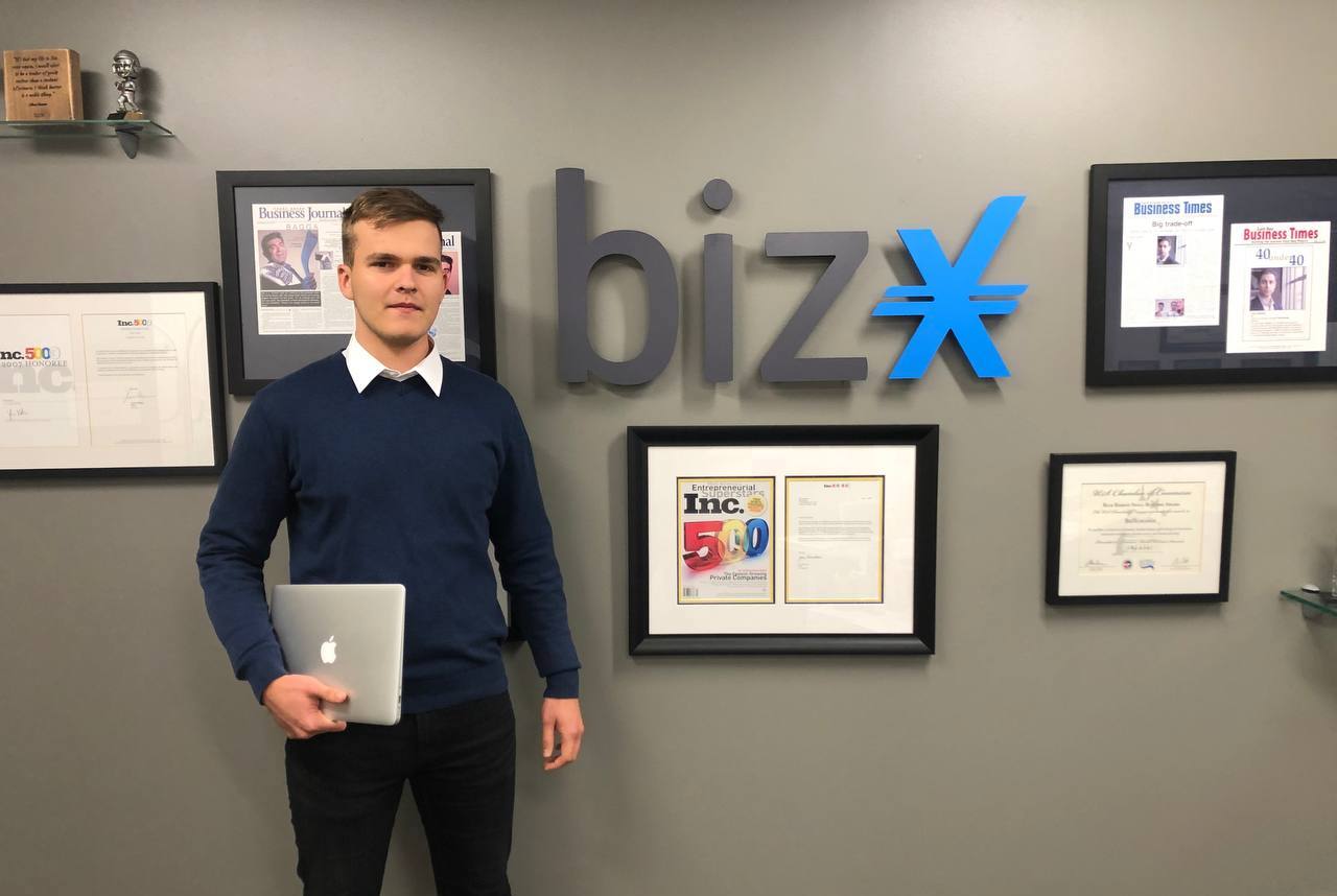 male wearing blue sweater standing in front of a grey wall with a biz x sign
