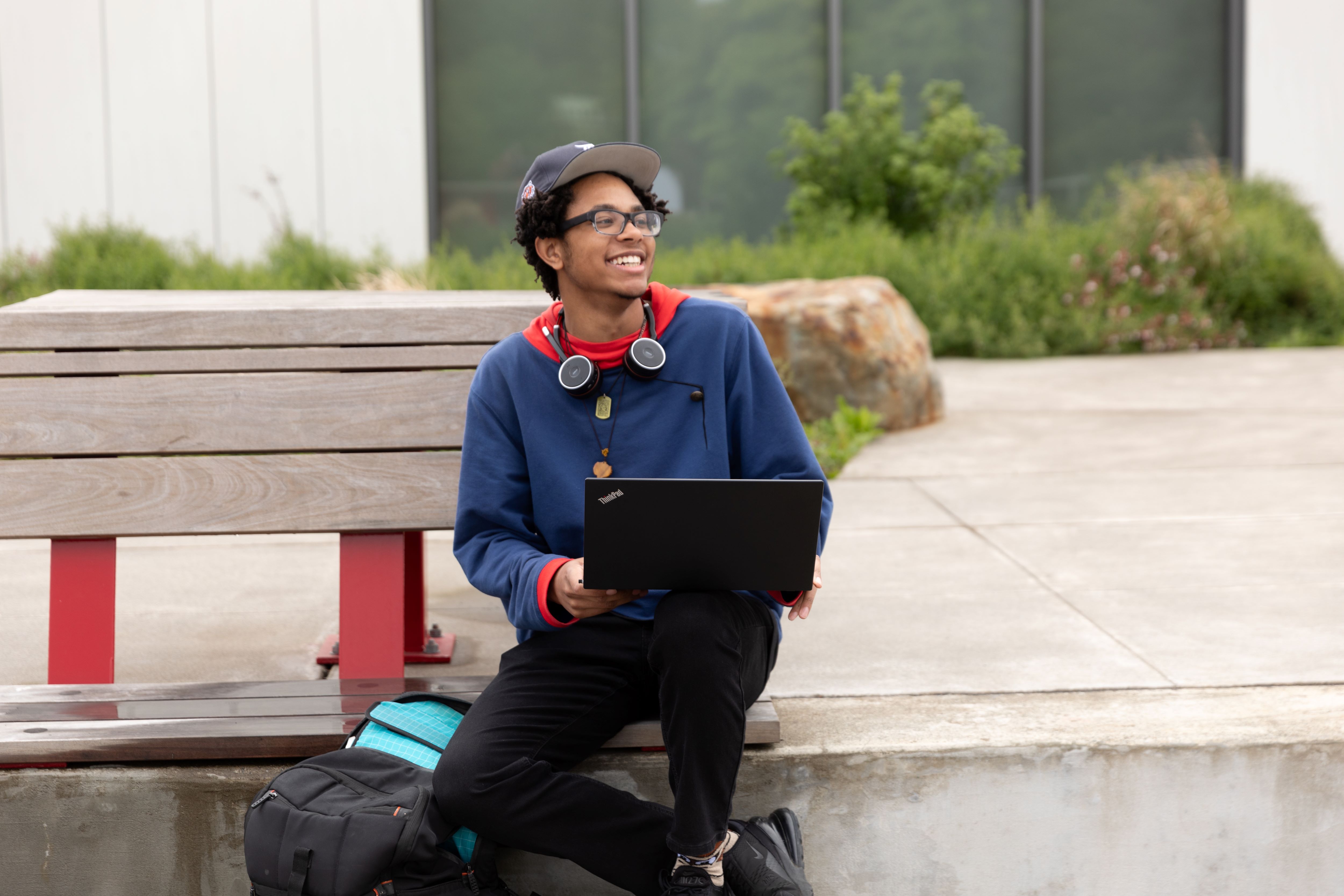 male wearing a blue shirt and holding a laptop and sitting on a bench outside