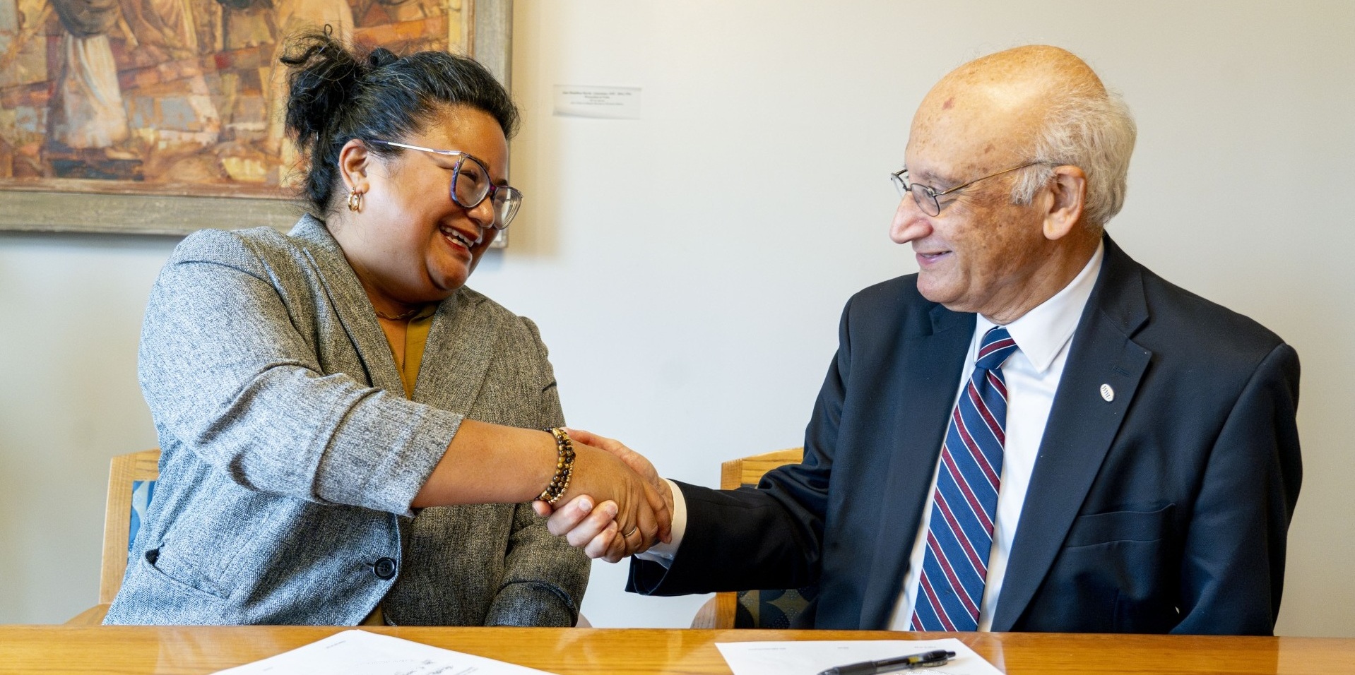 female and male smiling and shaking hands while sitting at a desk 