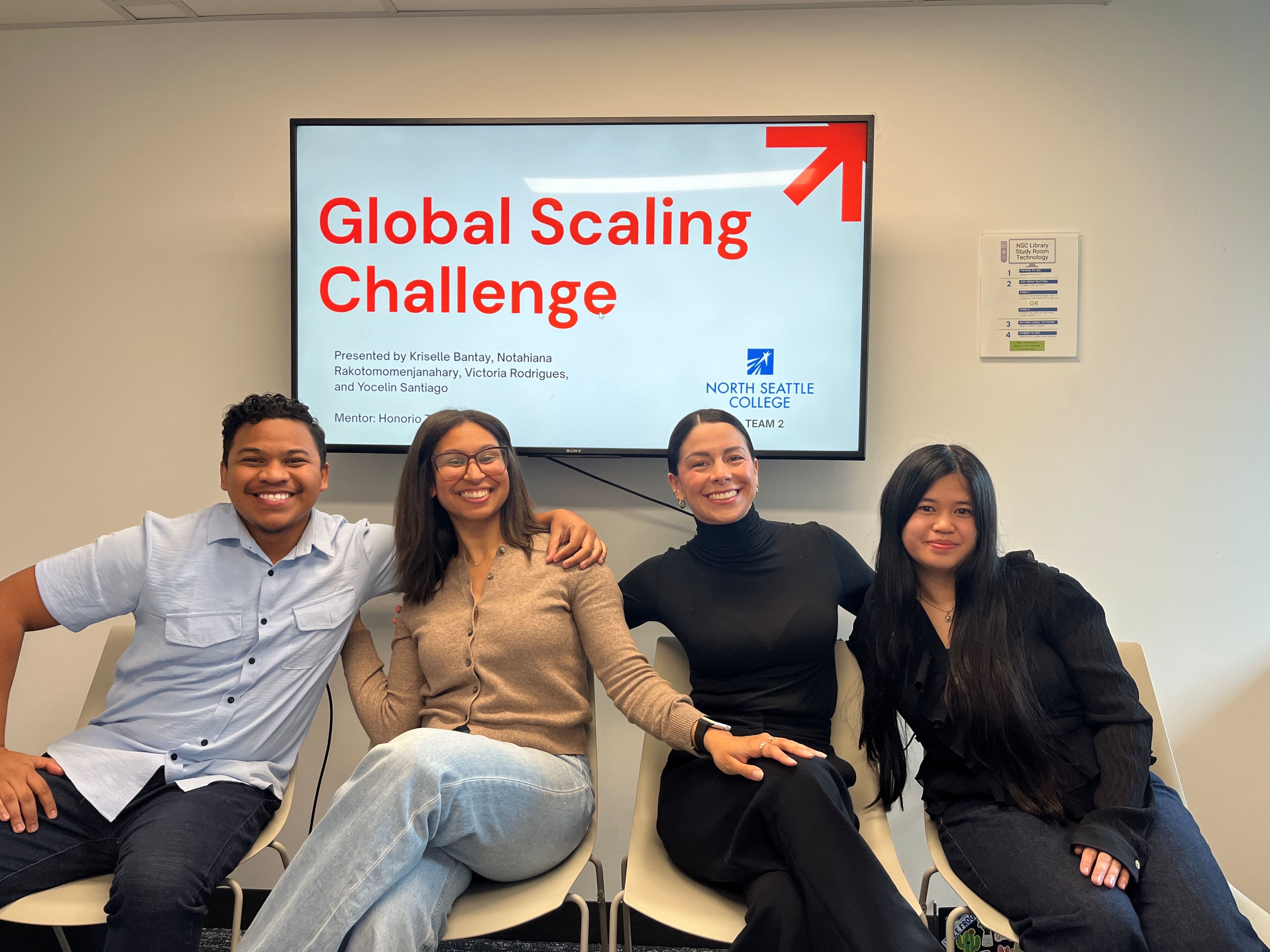 one male and three females sitting on chairs smiling with a digital monitor in the background that reads Global Scaling Challenge