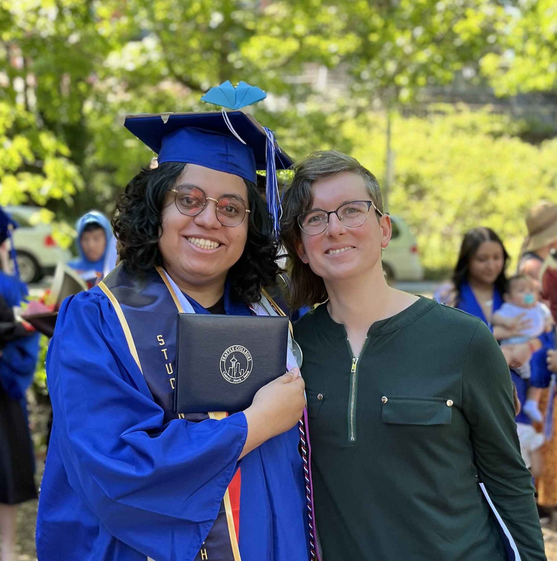 Female student with blue cap and gown holding a diploma smiling standing with another female outside 