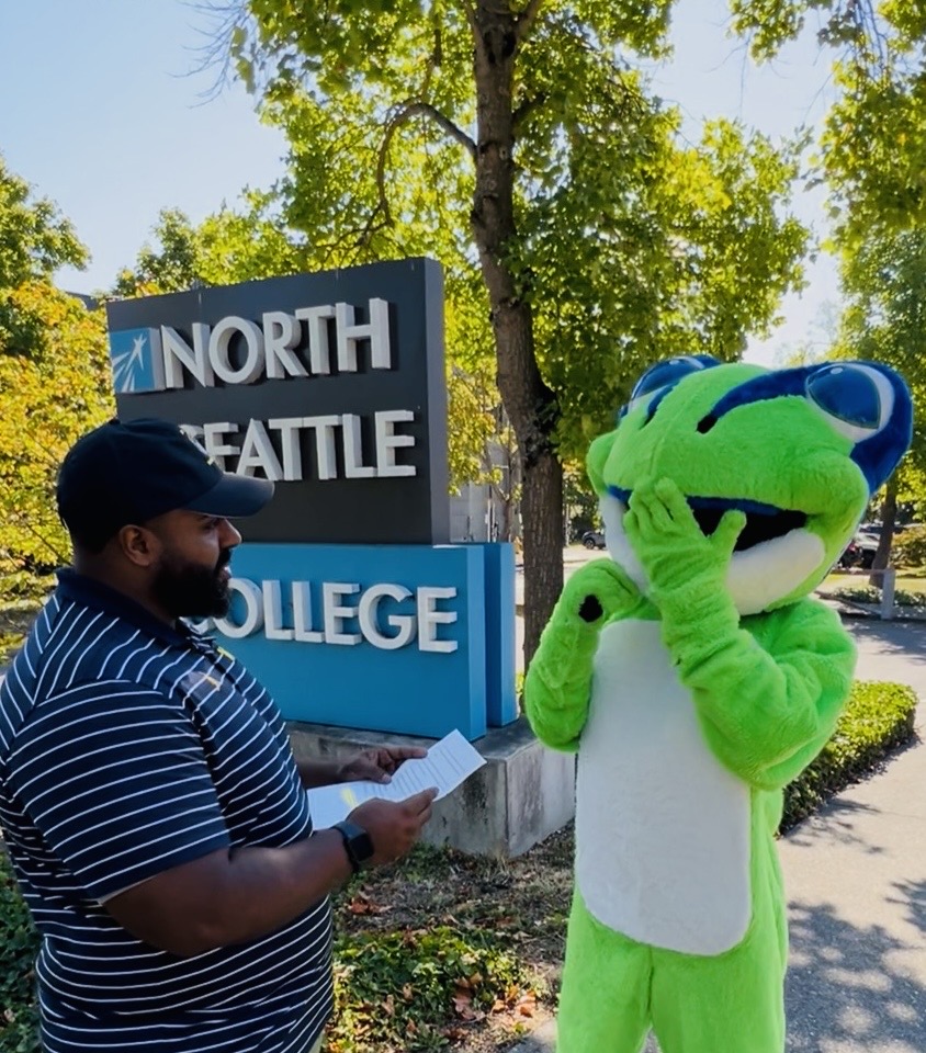 a frog mascot standing in front of a North Seattle College sign with trees in background