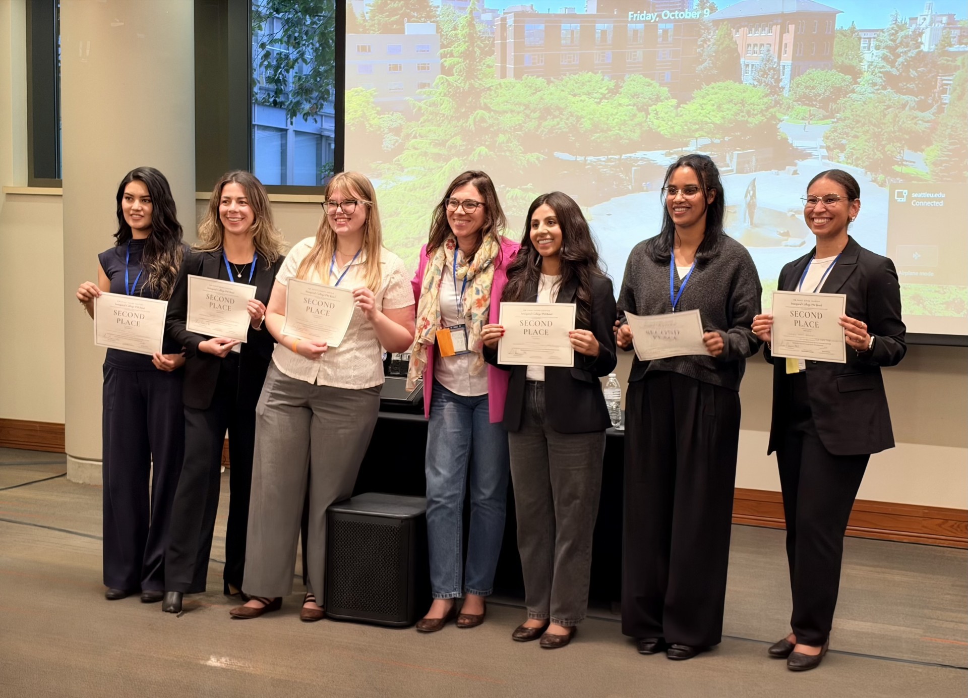 females wearng black business casual wear standing and smiling in front of a projector holding signs certificates that read second place 