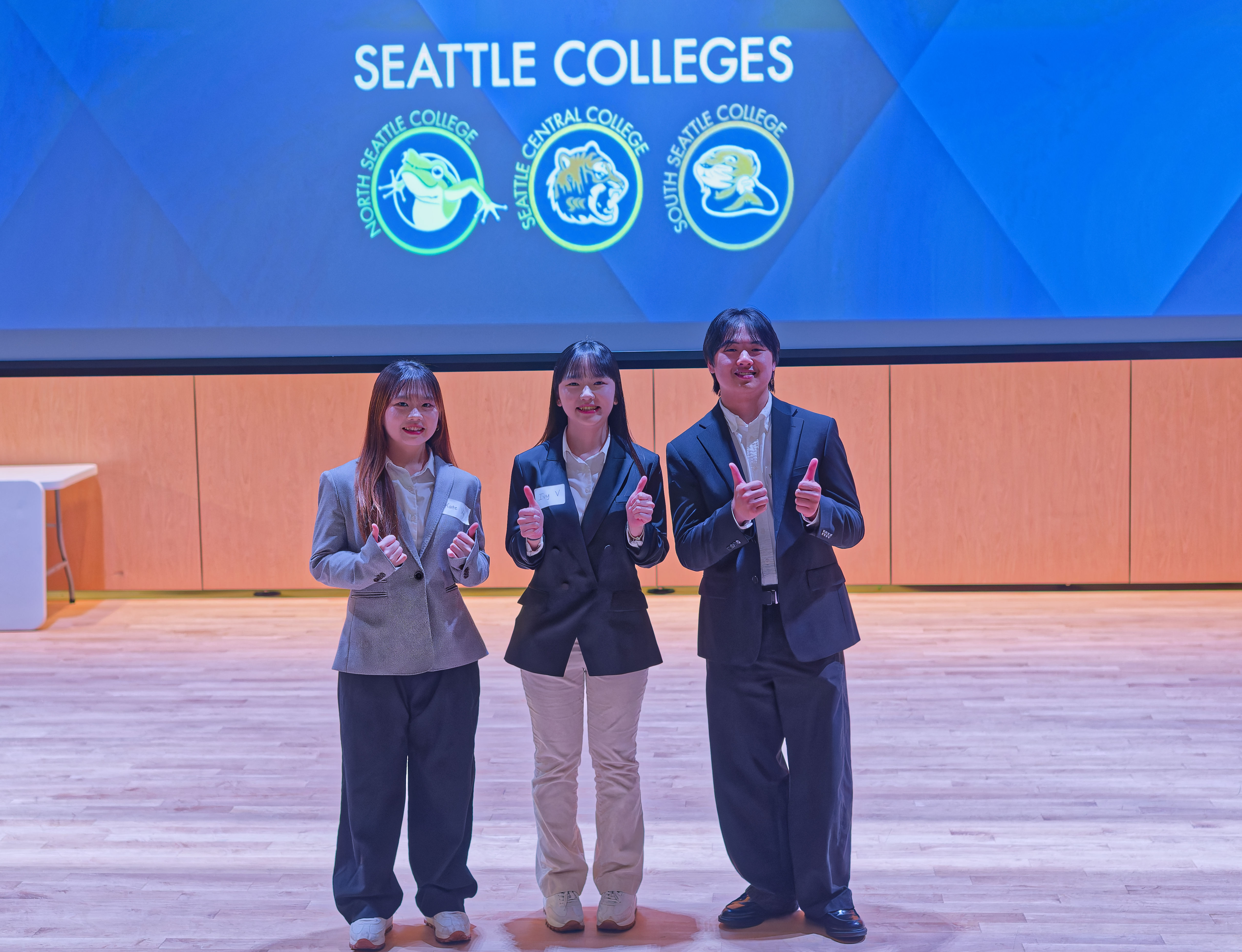 3 students standing on a stage in front of a blue background with the words Seattle Colleges wearing blazers holding thumbs up